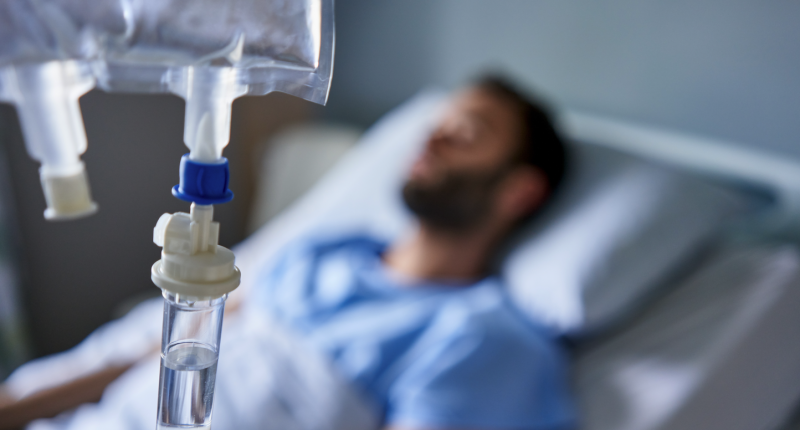 A man lies in a hospital bed receiving an IV drip.